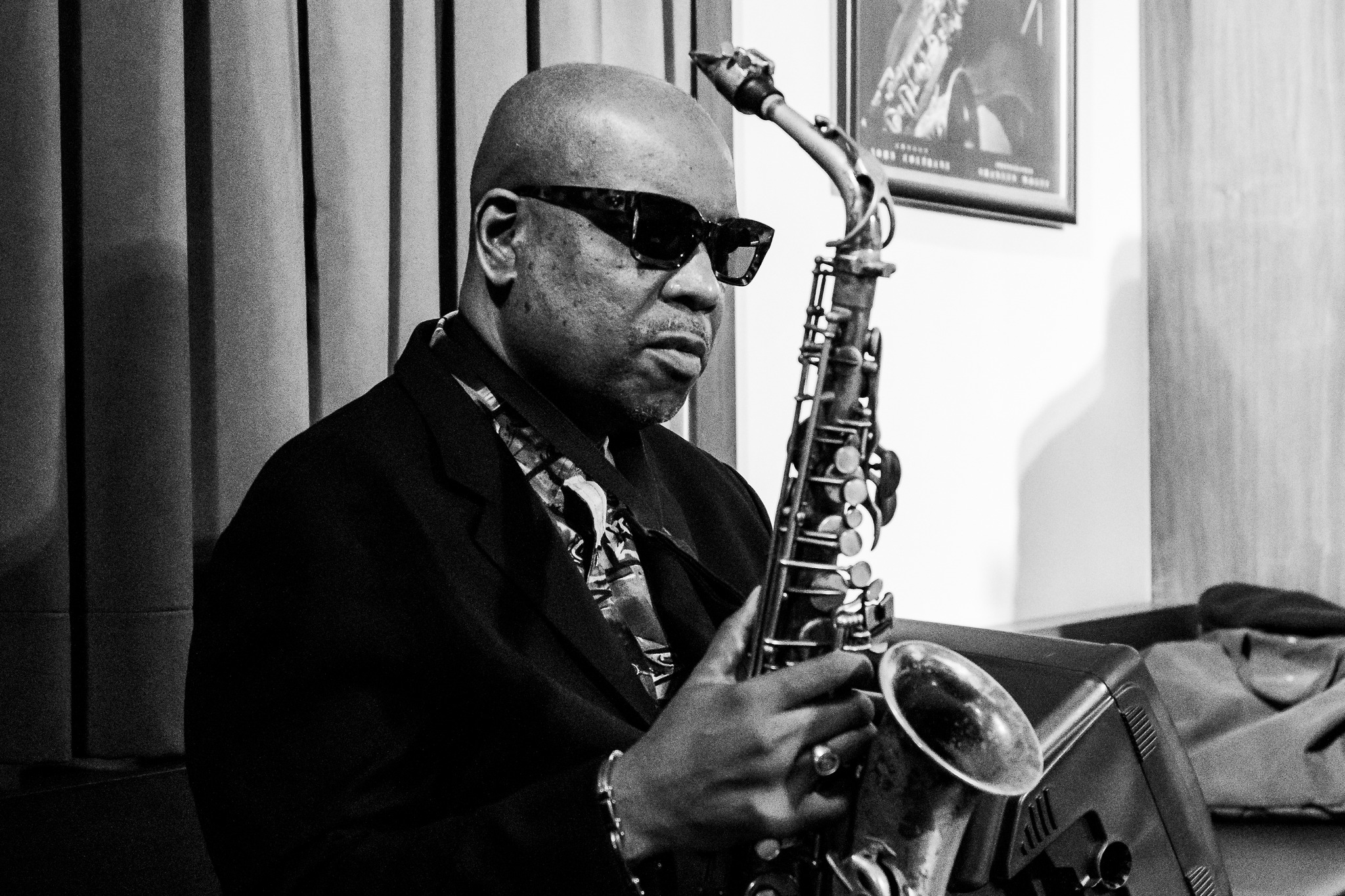 Black-and-white photo of a bald man wearing sunglasses, playing a saxophone in a recording studio.