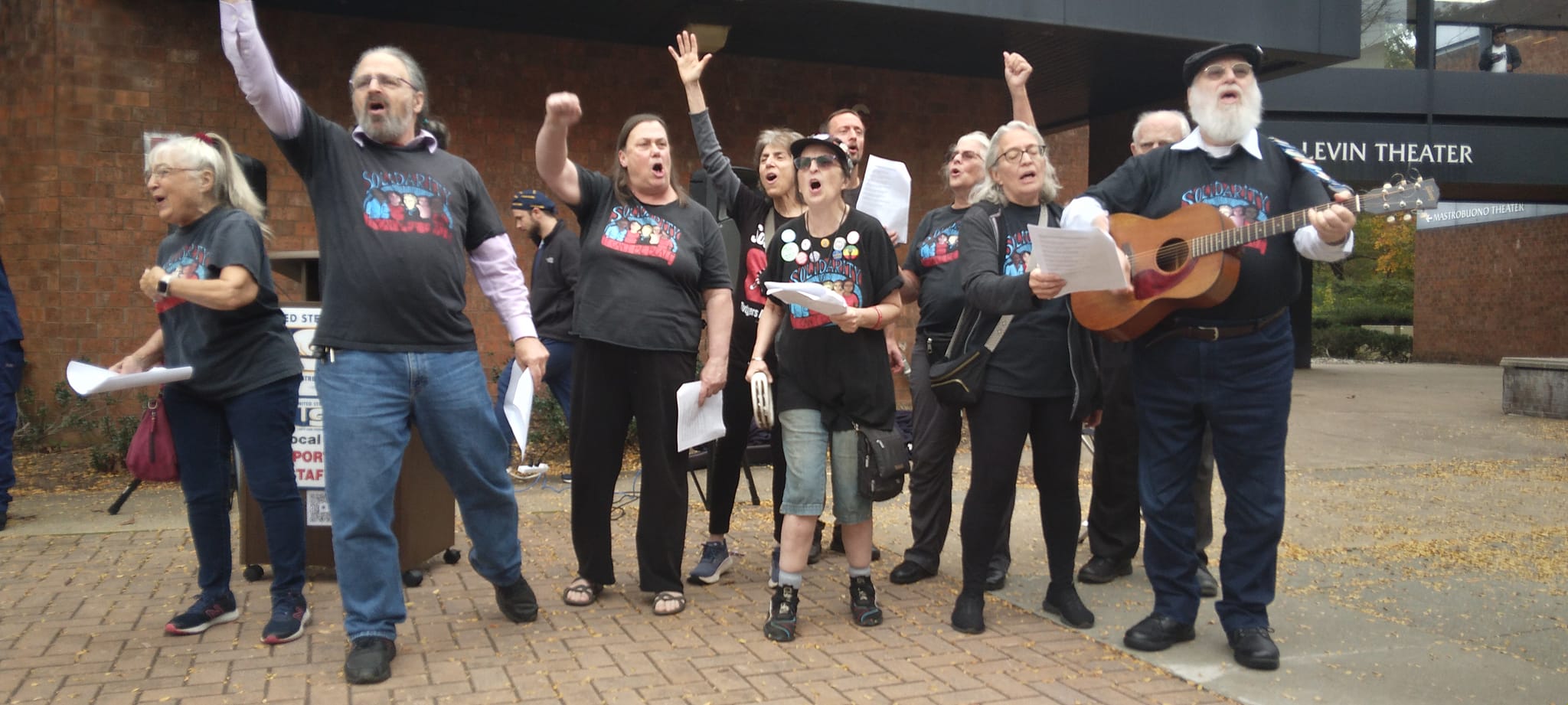 Group of people singing protest songs outside a brick building, with one man playing a guitar on the right.